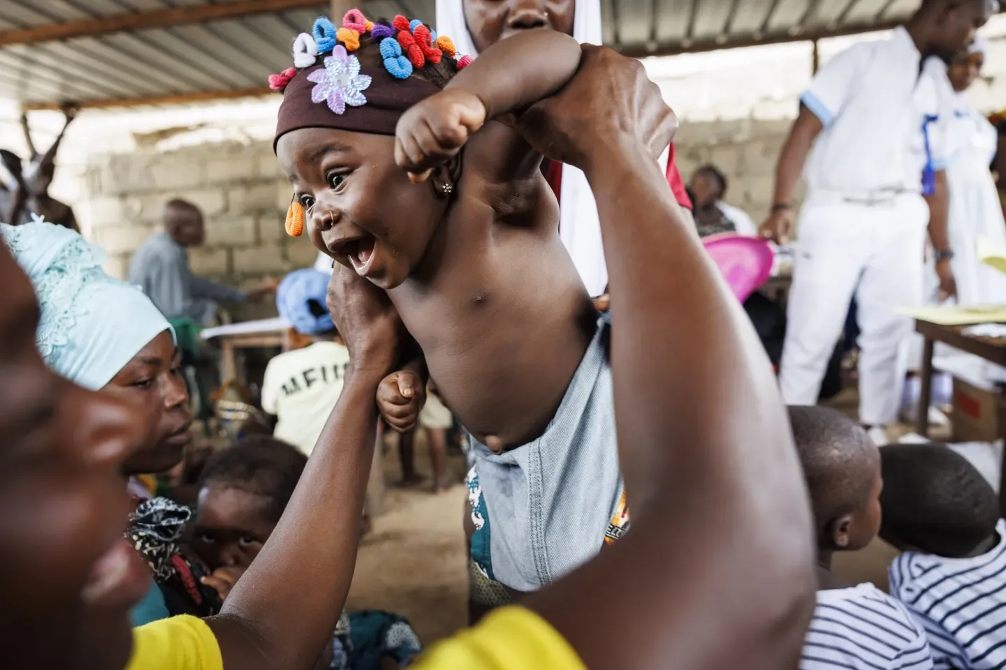 A joyful baby wearing a colorful headband is lifted in the air by an adult in a lively community setting.