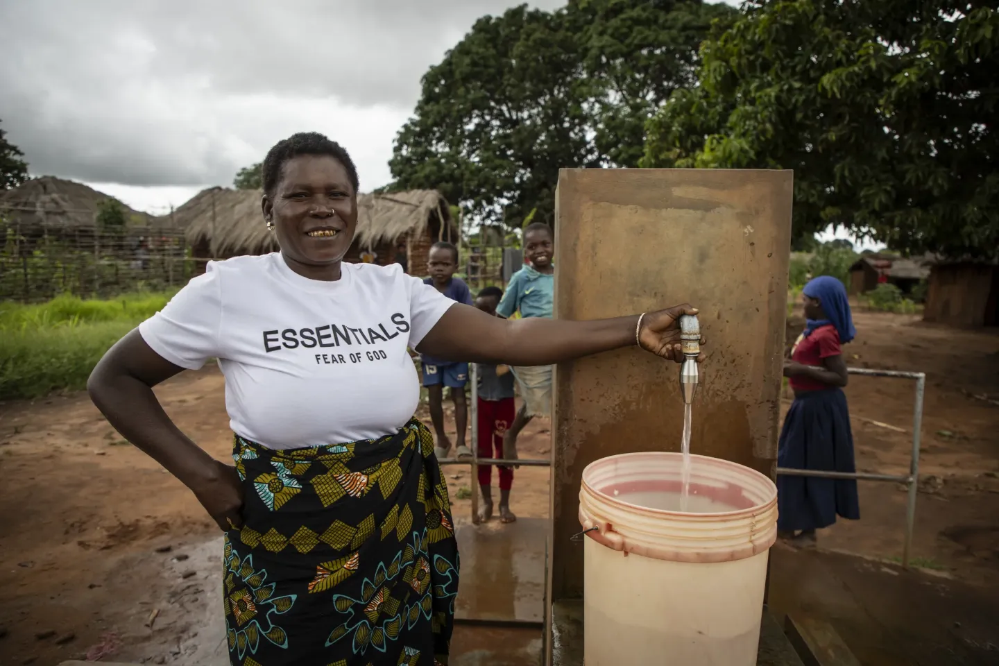 Woman filling a large container with clean water from a communal tap in a rural village setting with children and huts in the background.