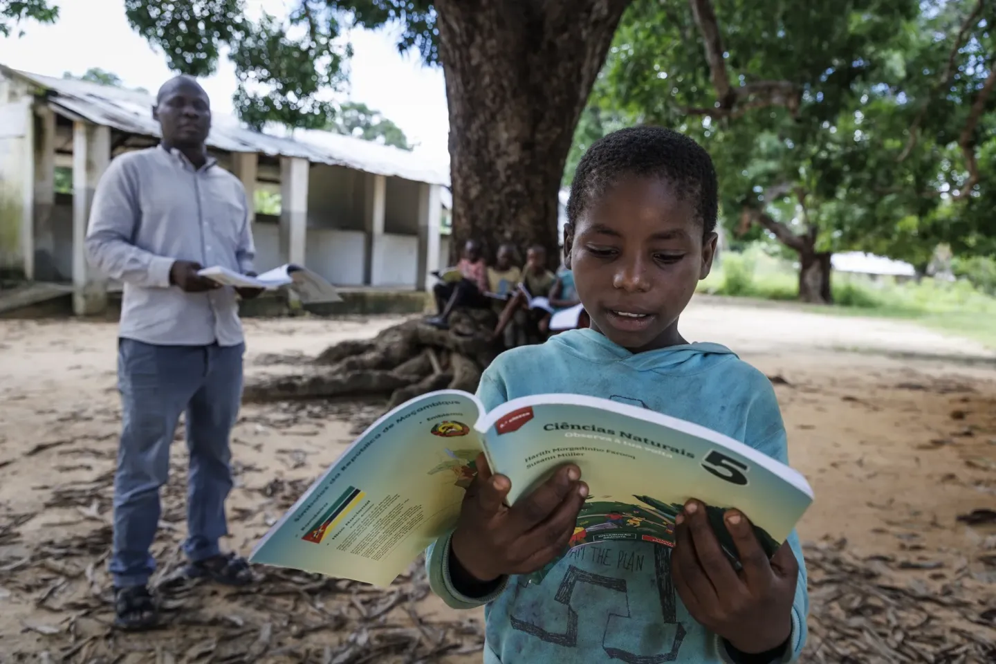 Young boy reading a colorful textbook outdoors under a large tree with a man holding papers nearby.
