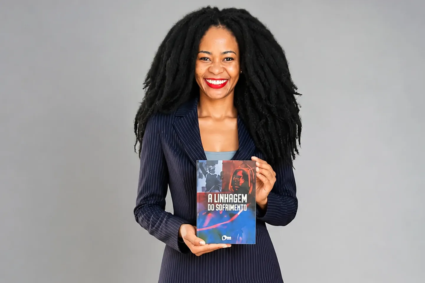 Mulher sorridente de blazer azul, cabelo crespo longo, segurando um livro colorido.