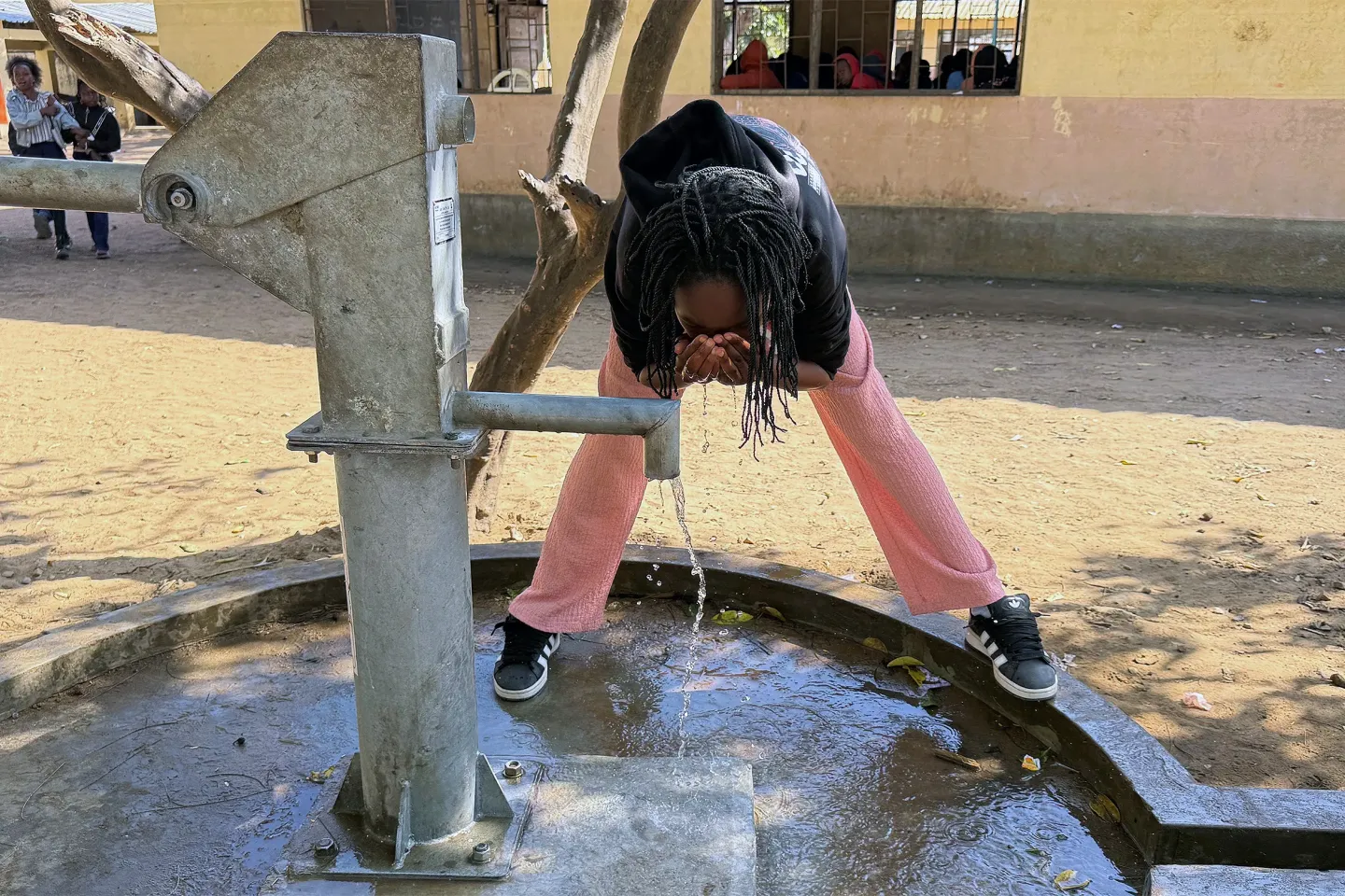 Pessoa bebe &aacute;gua em bomba manual ao ar livre, inclinada sobre o jato, de cal&ccedil;a rosa e moletom preto.