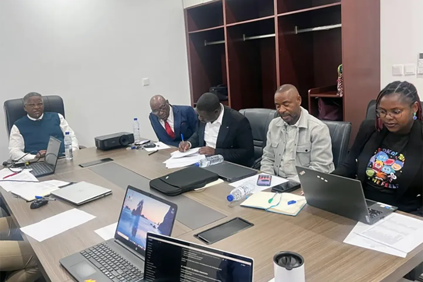 Reuni&atilde;o de trabalho com tr&ecirc;s pessoas em mesa de confer&ecirc;ncia, laptops abertos e anota&ccedil;&otilde;es.