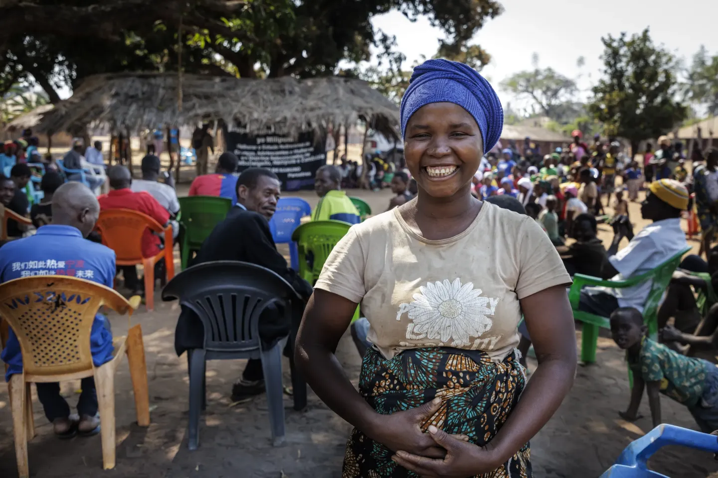 Mulher sorridente com turbante azul em frente a reuni&atilde;o comunit&aacute;ria ao ar livre, cadeiras coloridas e &aacute;rvores ao fundo.