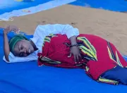 A performer, playing a girl in early marriage, lies still on a tarp during ASCHA's Theatre of the Oppressed performance in Horta-C, as another actor stands over her and the audience watches.