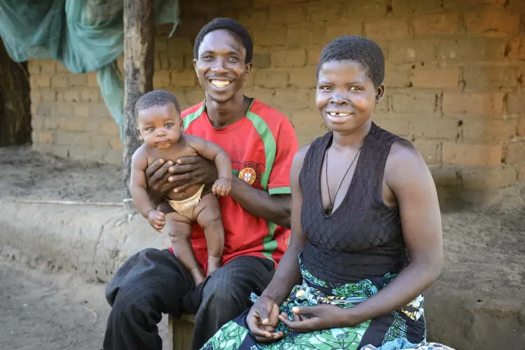 Uma fam&iacute;lia de tr&ecirc;s pessoas, sorridente, sentada &agrave; porta de uma casa r&uacute;stica de tijolo; o pai segura um beb&eacute; e a m&atilde;e est&aacute; sentada ao lado deles.