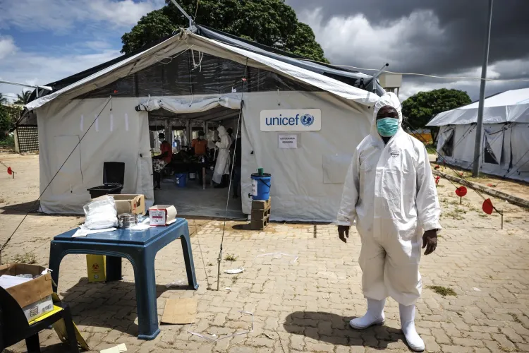 Profissional de sa&uacute;de com roupa protetora diante de tenda da UNICEF, mesa com suprimentos e c&eacute;u nublado.