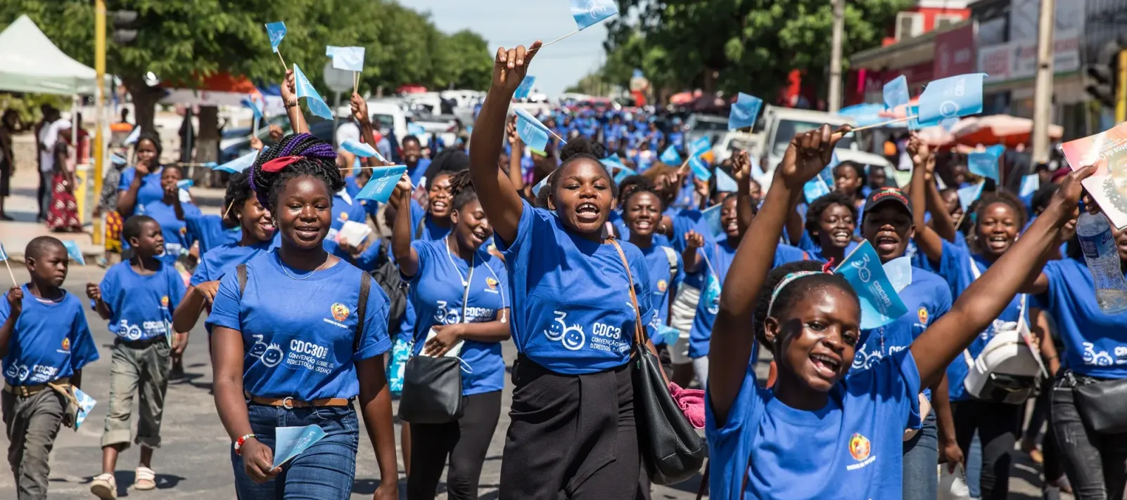 A large group of joyful people wearing matching blue shirts march down a street waving small blue flags during a daytime event.