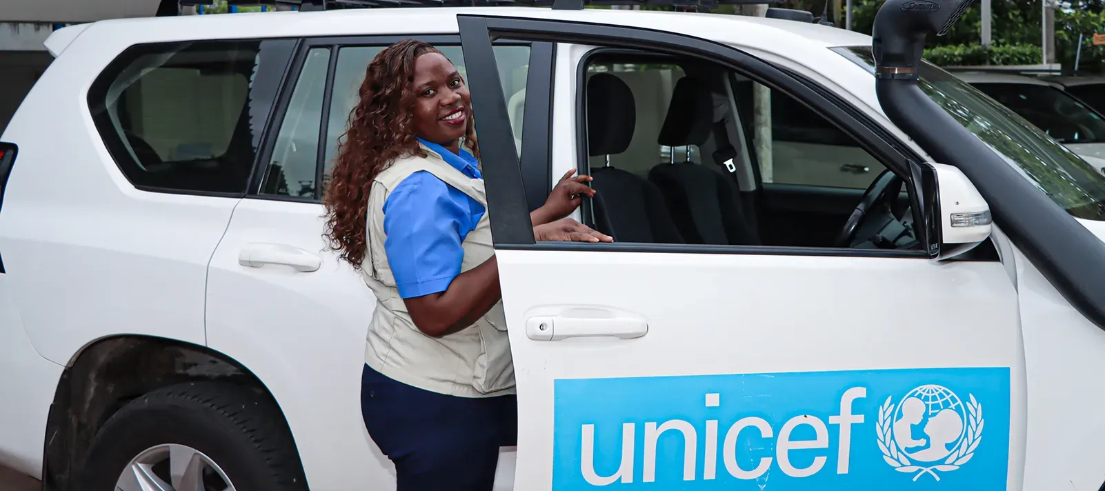 Mulher sorridente abre a porta de um carro branco com o logotipo da UNICEF.
