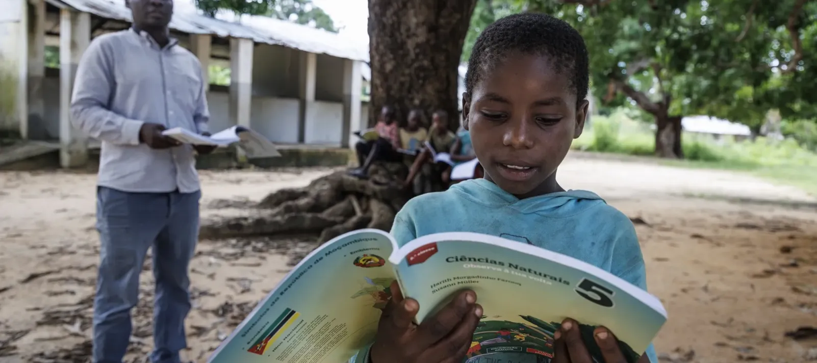 Young boy reading a colorful school textbook outdoors under a large tree with a teacher nearby holding papers.