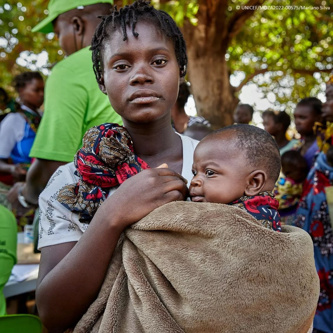 A mother carrying her baby child in a blanket wrapped around her chest looks at the camera. The mother stands in front of a group of mothers with their babies queuing to receive the child grant support.