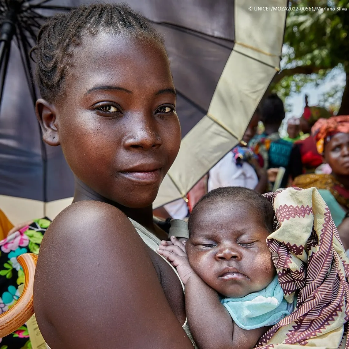 A mother looks at the camera while holding her sleeping baby next to her chest.