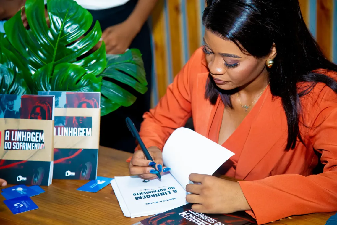 Mulher de blazer laranja autografa livro em mesa, com exemplar exposto ao lado e planta verde pr&oacute;xima.