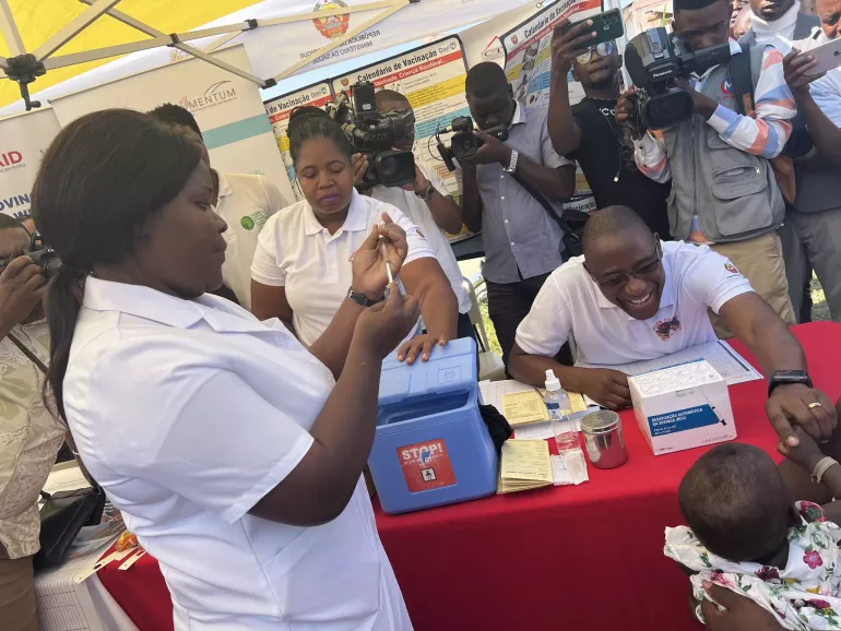 A health technician prepares a malaria vaccine while the Minister of Health plays with a child on his mother's lap.