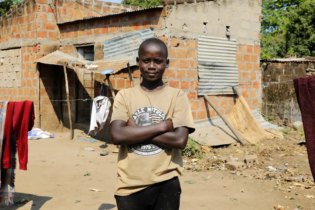 Young boy stands confidently with arms crossed in front of a rustic brick and metal sheet house in a rural setting.