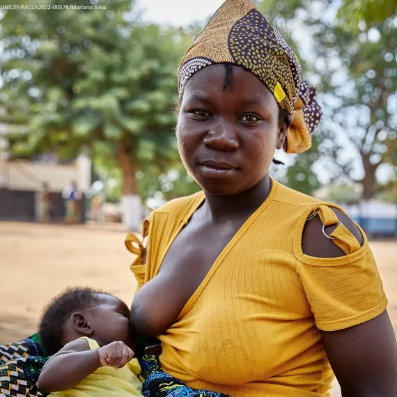 A mother seated on the floor wearing a yellow shirt looks at the camera while breastfeeding her baby resting on her lap.