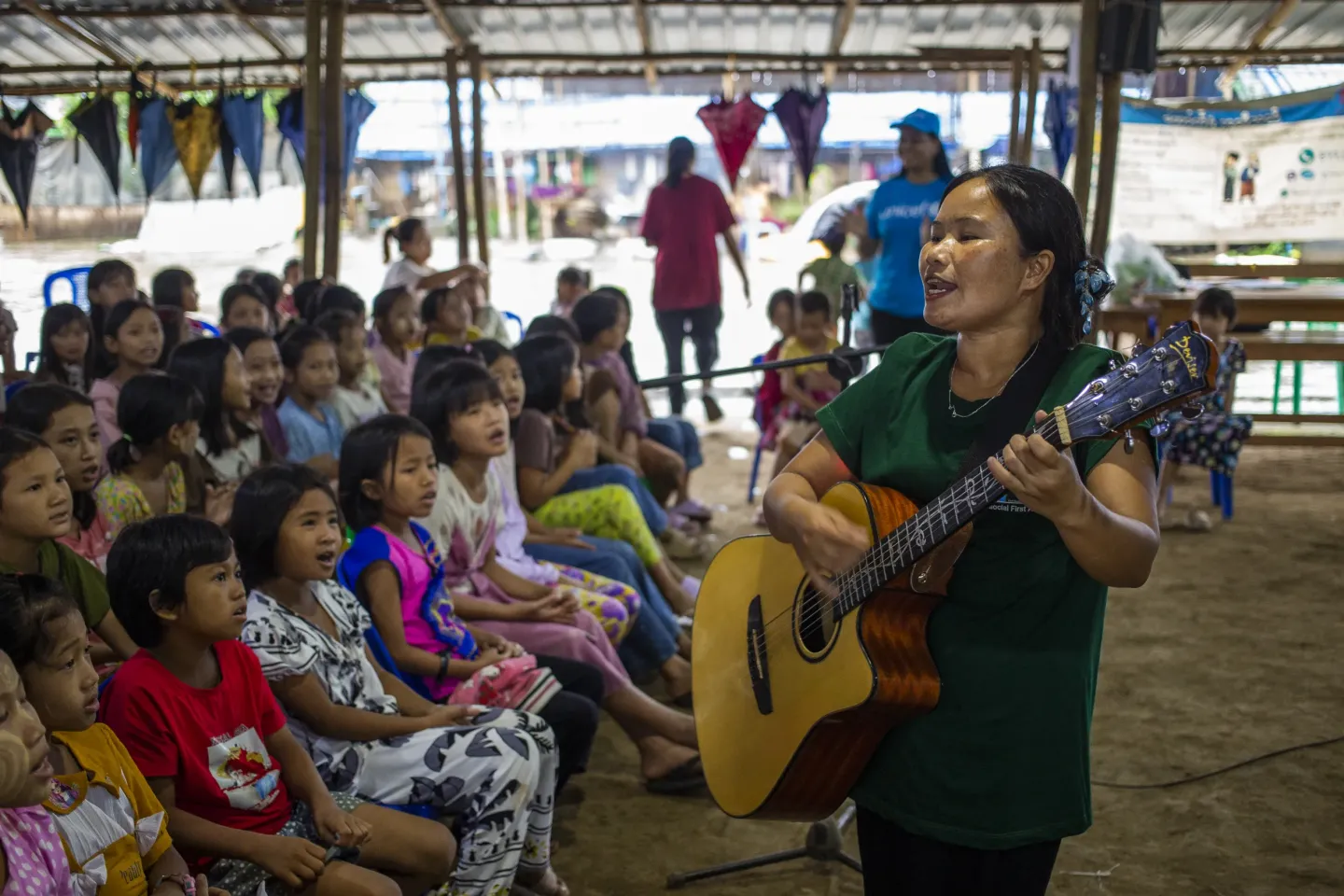 A woman singing with a guitar in front of a group of young children inside a big tent. 