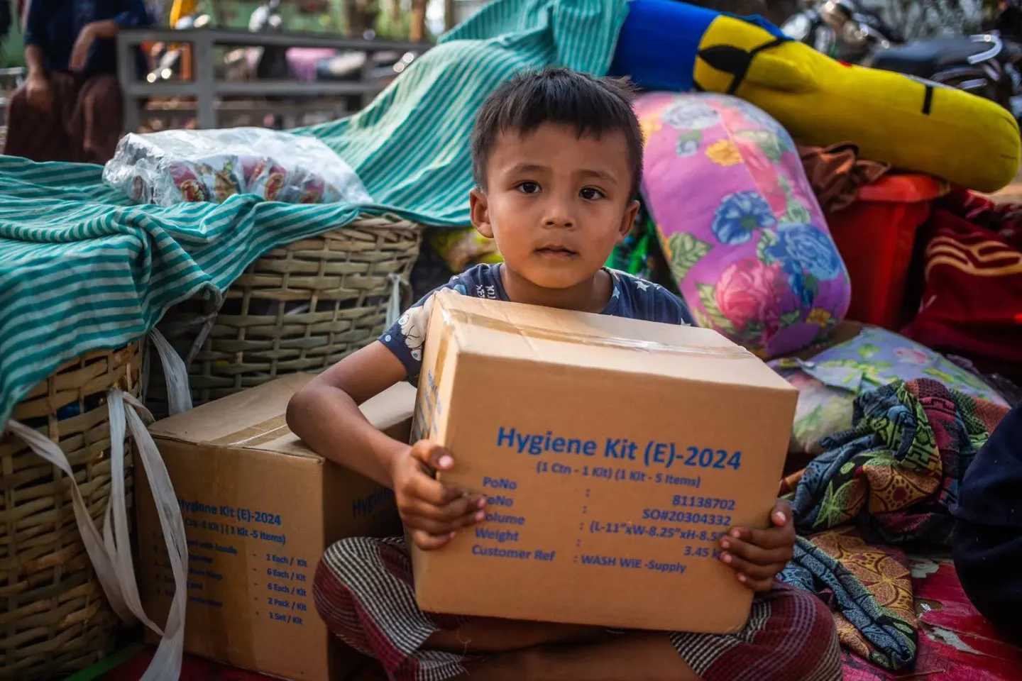 A young boy sits among household belongings and bedding, holding a UNICEF hygiene kit box during a humanitarian assistance distribution.