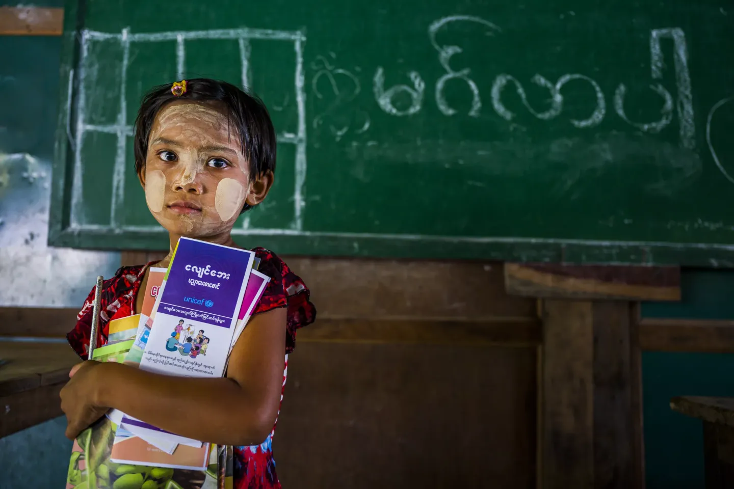 A young girl holding earthquake safety pamphlets in front of a black board inside a classroom. 