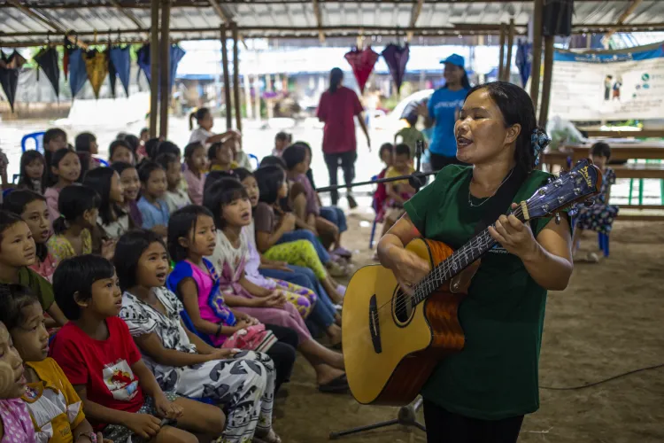 A woman singing with a guitar in front of a group of young children inside a big tent. 