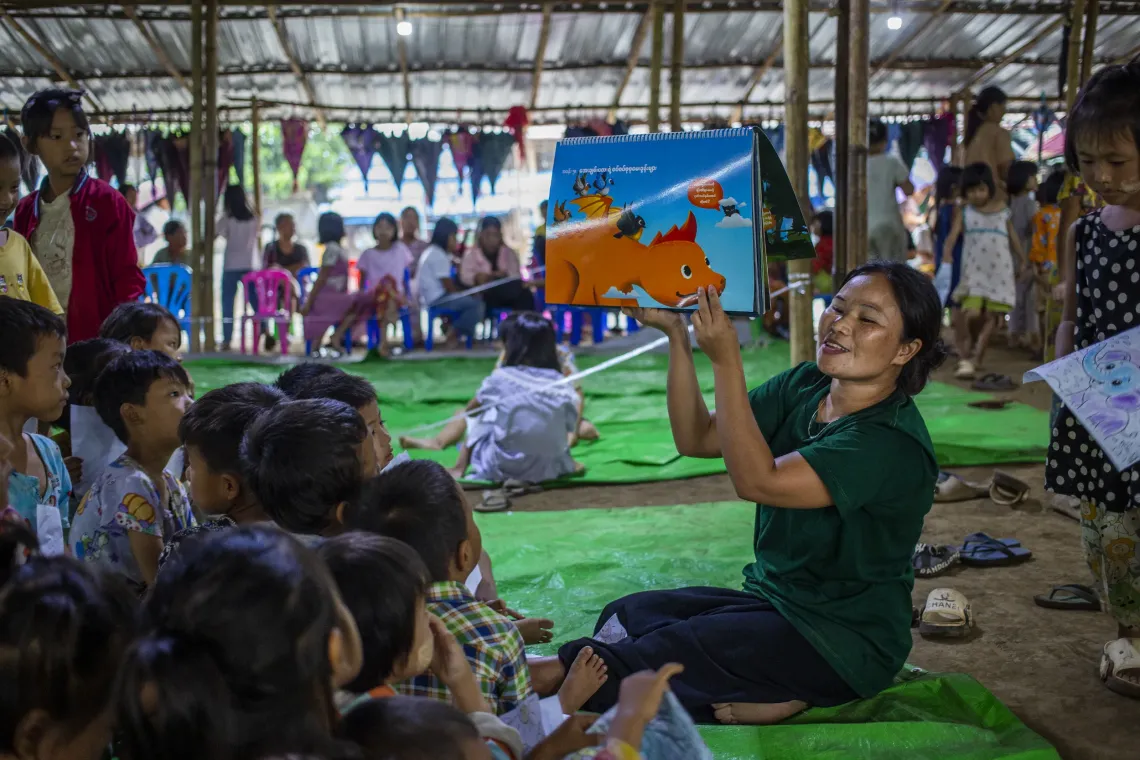 A woman uses an illustrated storybook to tell stories to a group of young children in side a tent. 