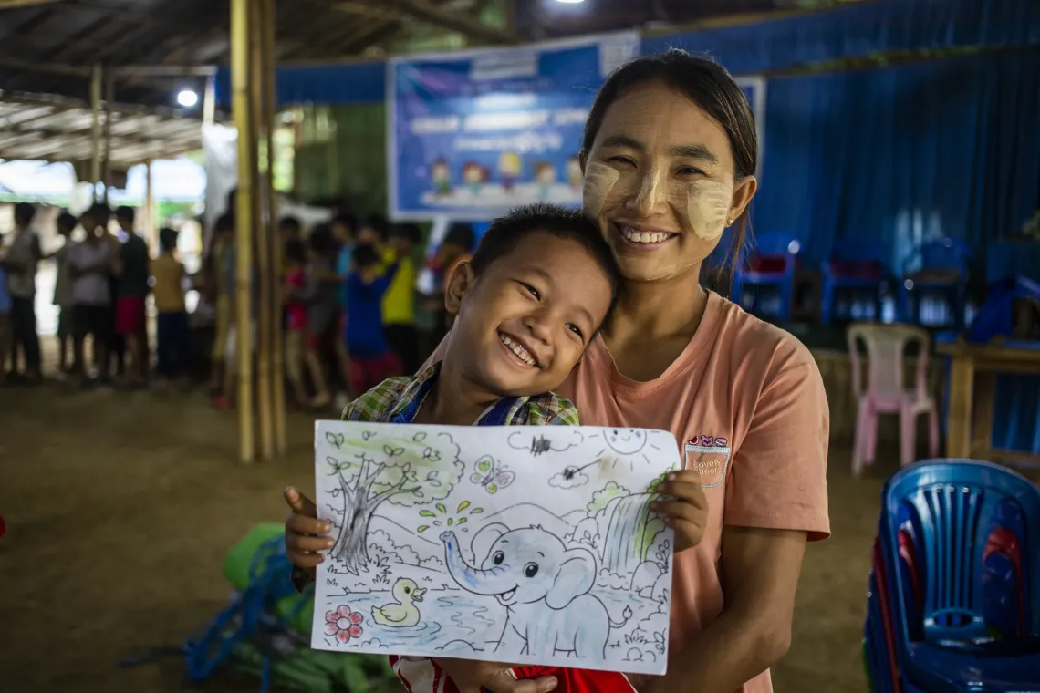 A mother and her son smiling while the boy holds a piece of paper showing what he colored during a child friendly space activity. 