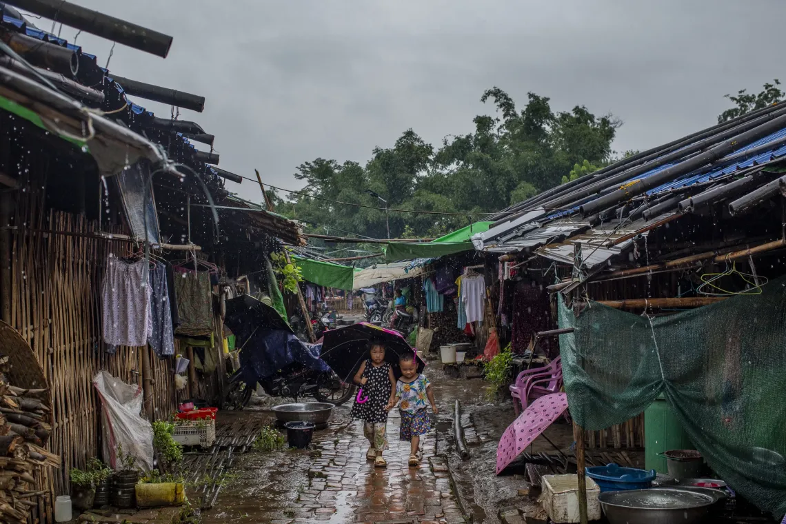 Two young children walk along a muddy path between rows of temporary shelters in a displacement camp, during the monsoon rain.