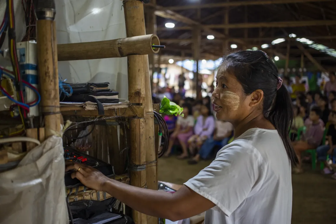 A woman uses audio control panel while there are a group of children in the background singing. 