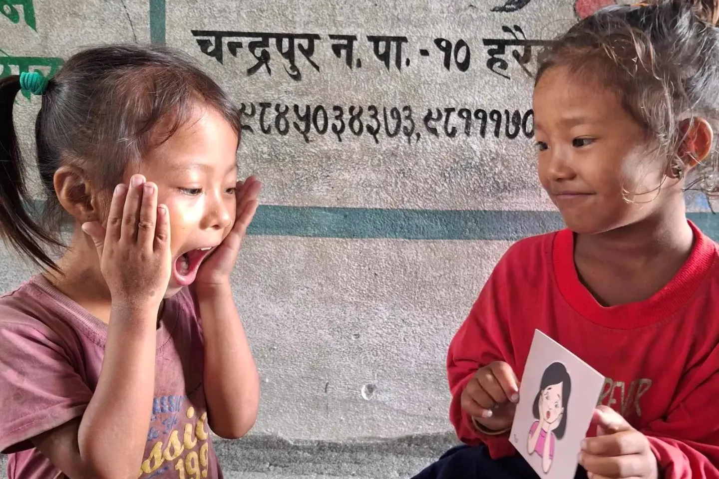 Two small girls sitting in the classroom while one of them is mimicking the emotion shown by the next girl.