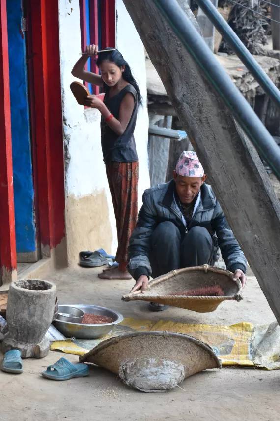 Jhim Bahadur Bhandari (right) sifts through a batch of millet as his youngest daughter Sarina (left) gets ready for school on the porch of their home in Bam Badha, Mugu District. 
