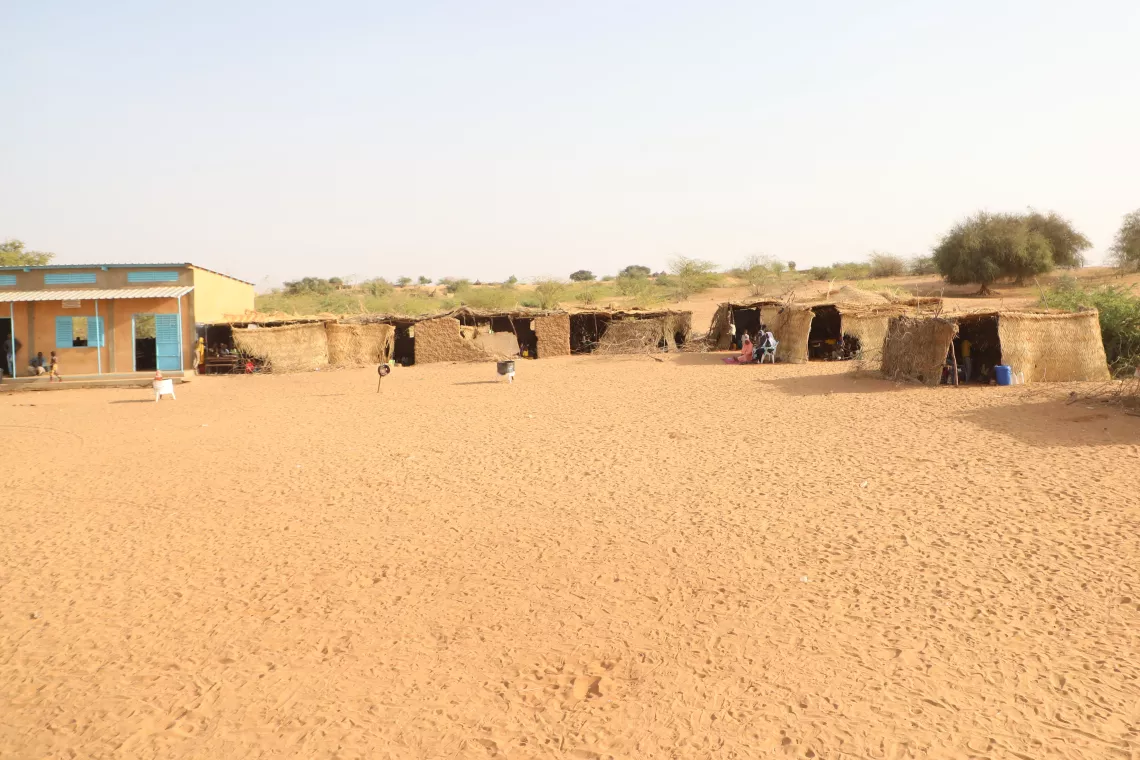 A view of several classrooms constructed of straw next to a schoolhouse built of bricks with two classrooms.