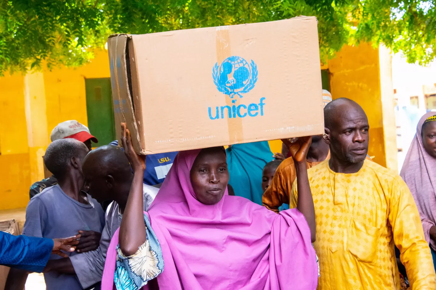 A women wearing a purple hijab holding a cardboard box with the UNICEF logo on it, on her head