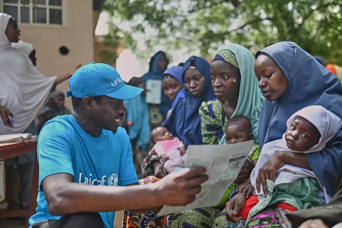  A man dressed in blue speaking to women who are holding children.