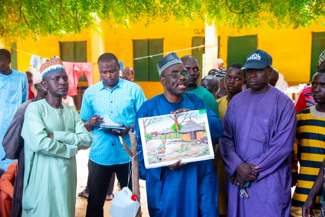 A group of men standing in a community while one of them speaks holding a drawing
