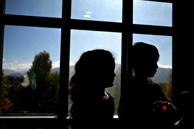 Two children stand in front of a window. 