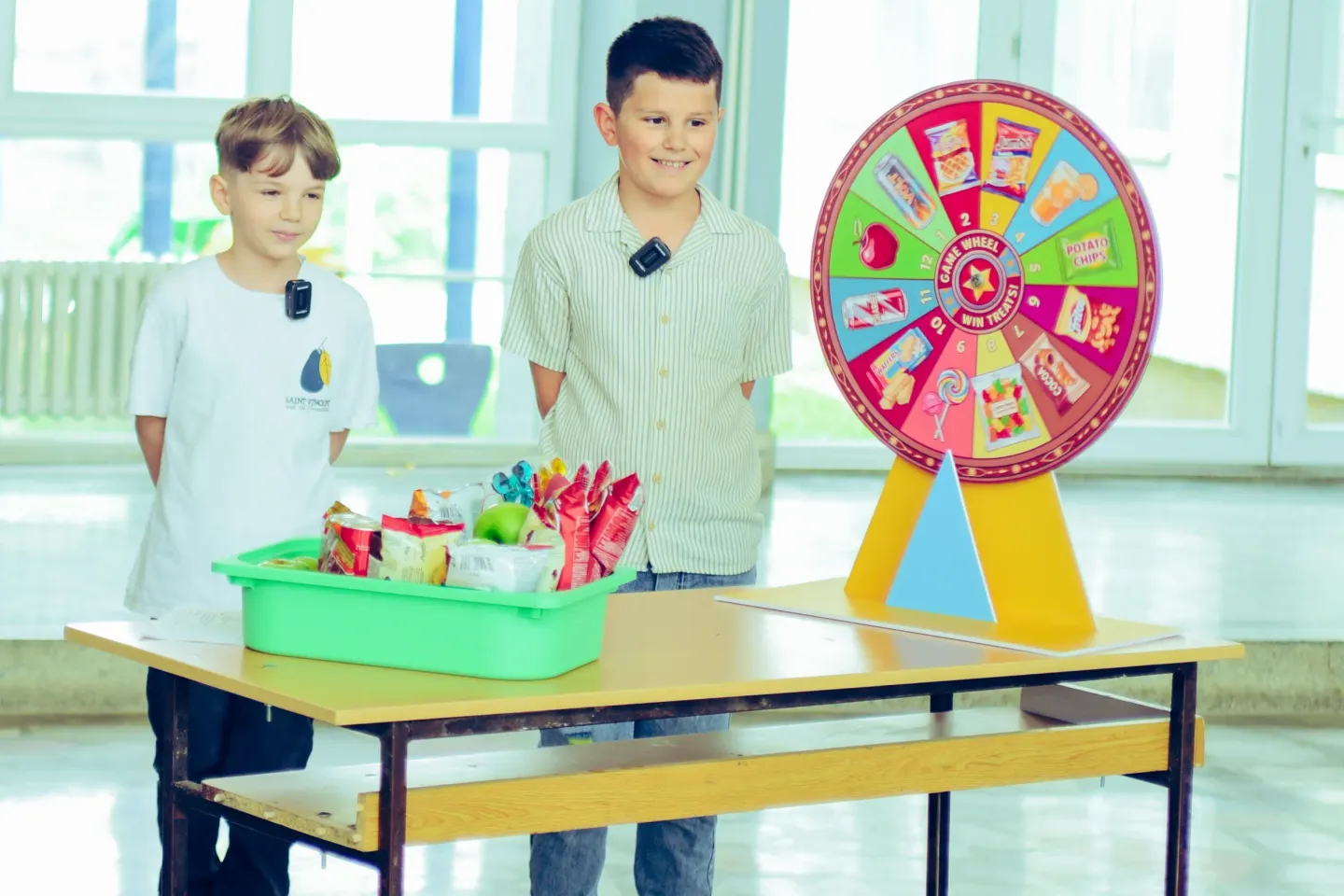 Two boys in school hallway standing beside a desk with a basket full of junk food and spinning wheel