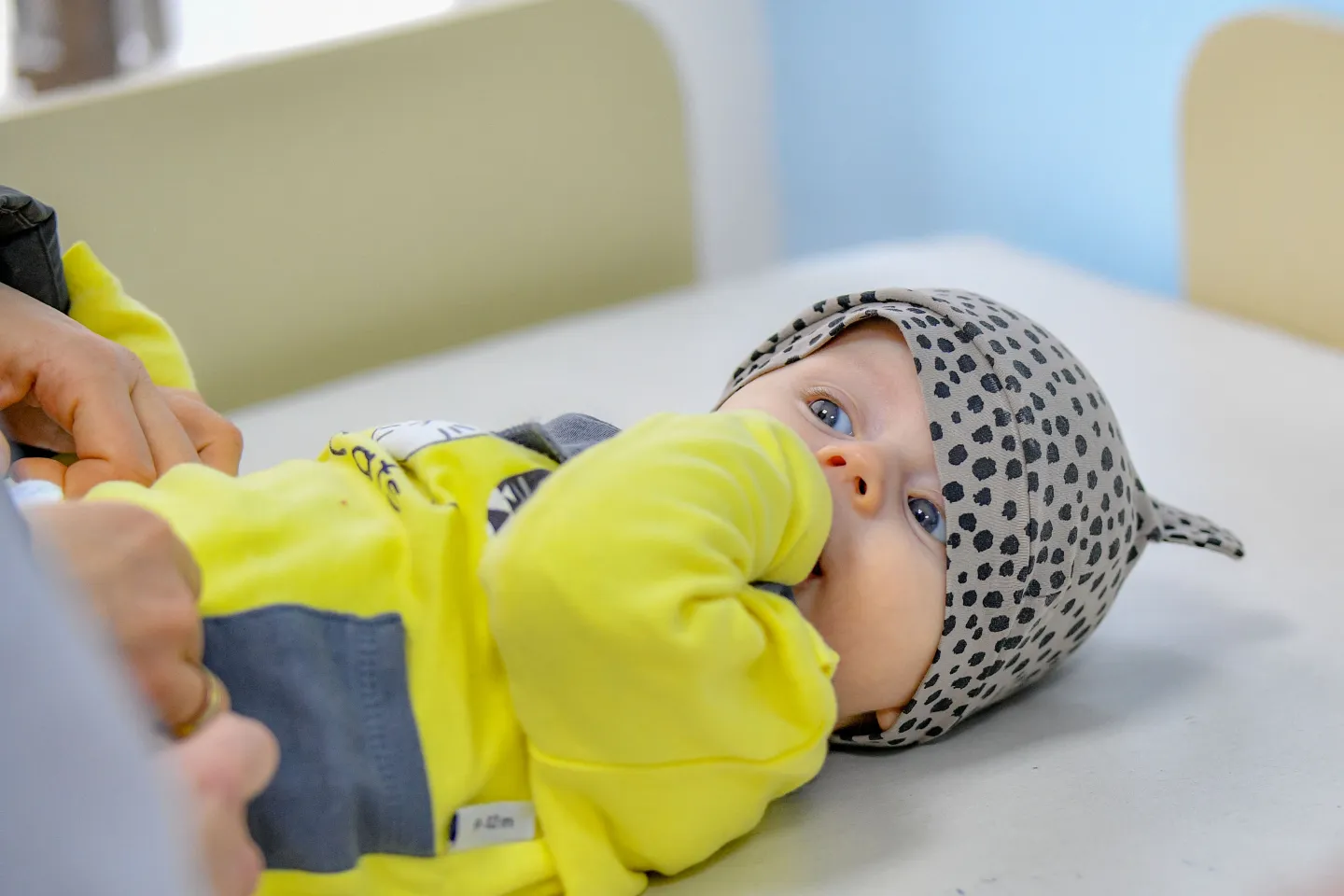 Baby boy in a yellow shirt sucking his fingers and looking at the nurse as she is preparing him for vaccination UNICEFMK