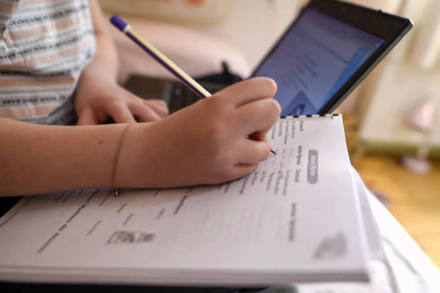 Child's hand holding a pen and taking notes with laptop open nearby