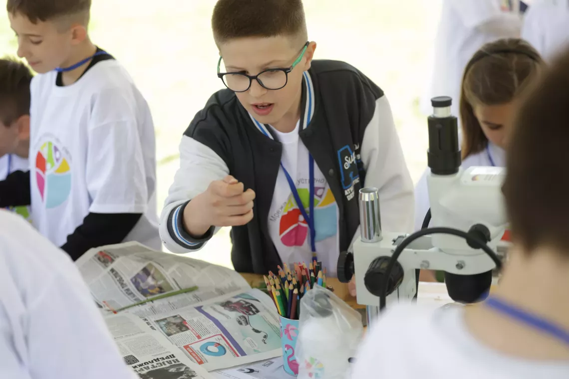 Boy wearing glasses and a black and white jacket looks closely at a plant during an experiment organized within the Climate Action event in Skopje