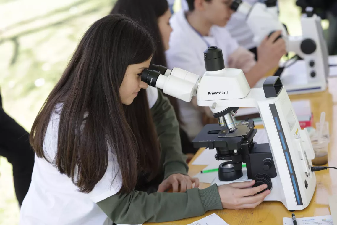 Girl with long brown hair and white t-shirt looks through a microscope during the experiential learning workshop at the Climate Action event in the park