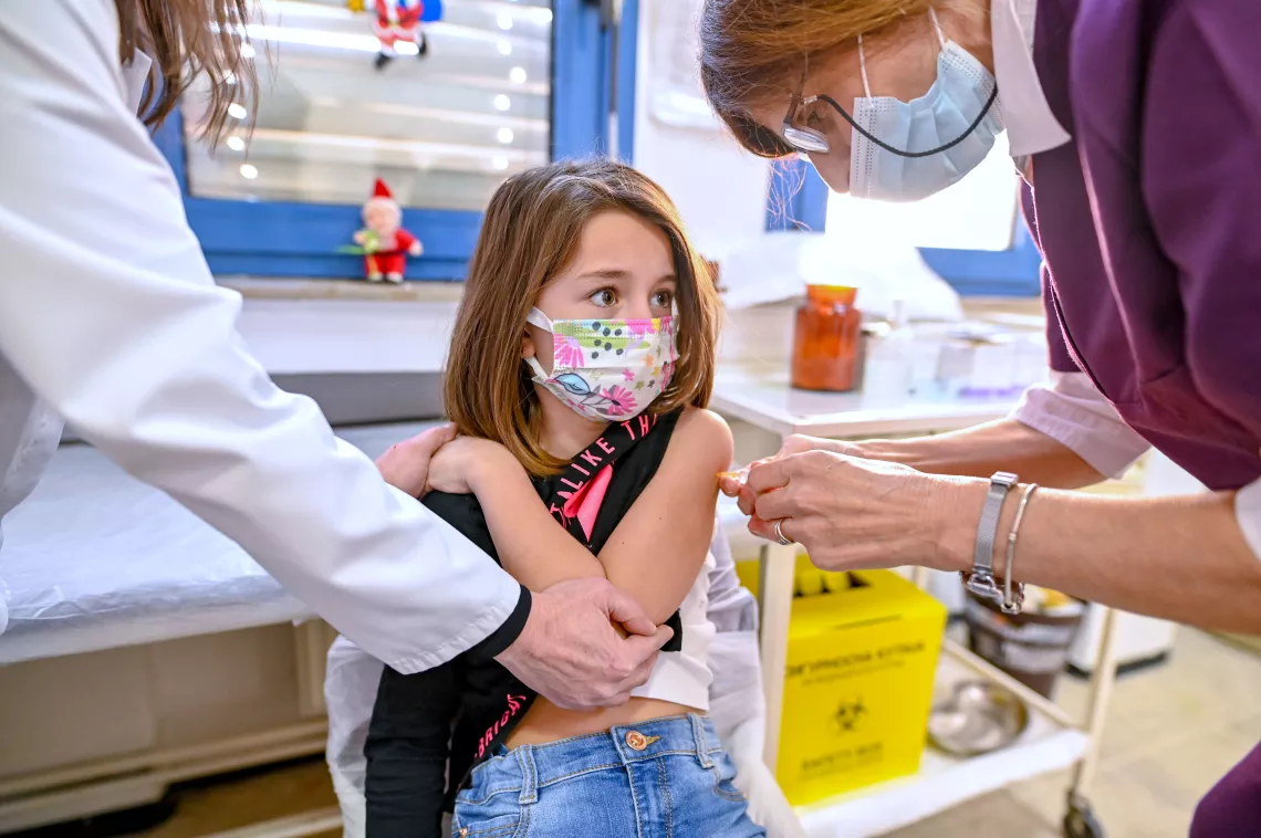 Nurse administering a vaccine to a young girl, while her pediatrician is comforting her and holding her hand