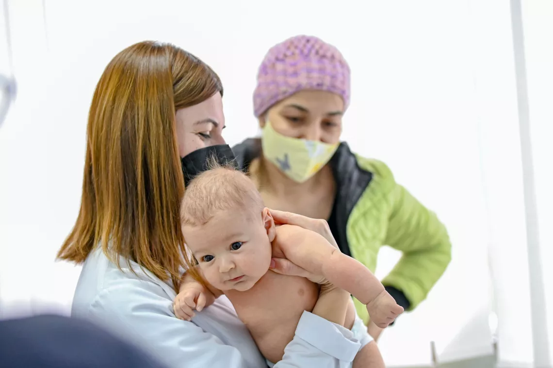 Doctor holding a baby and preparing for administrating a vaccine, while it's mother is observing in the background