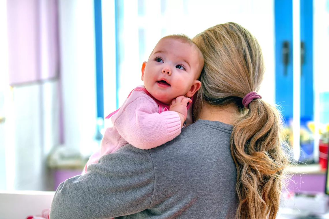 Mother turned with her back to the camera is holding a baby looking up amused 