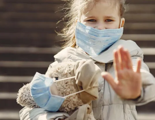 A girl wearing a mask, holding a teddy bear also wearing a mask