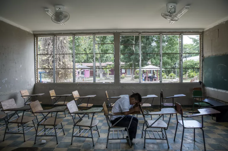A child sitting alone in a classroom with his head leaned on the chair in front of him