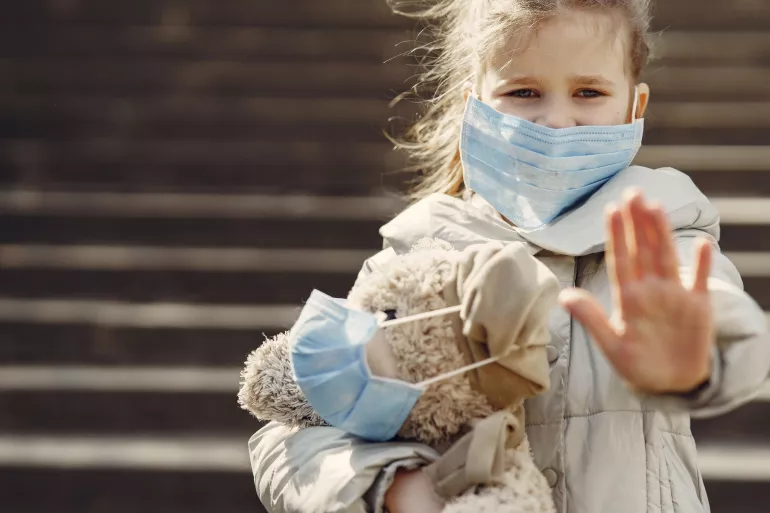 A girl wearing a mask, holding a teddy bear also wearing a mask