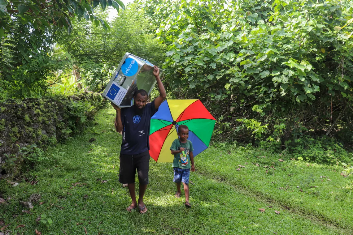 Michael Birikai, community volunteer, and his young son on their way to the nakamal to set up for the planned psychosocial support activities with Olapa Village children as well as those who made the journey from neighboring communities. 