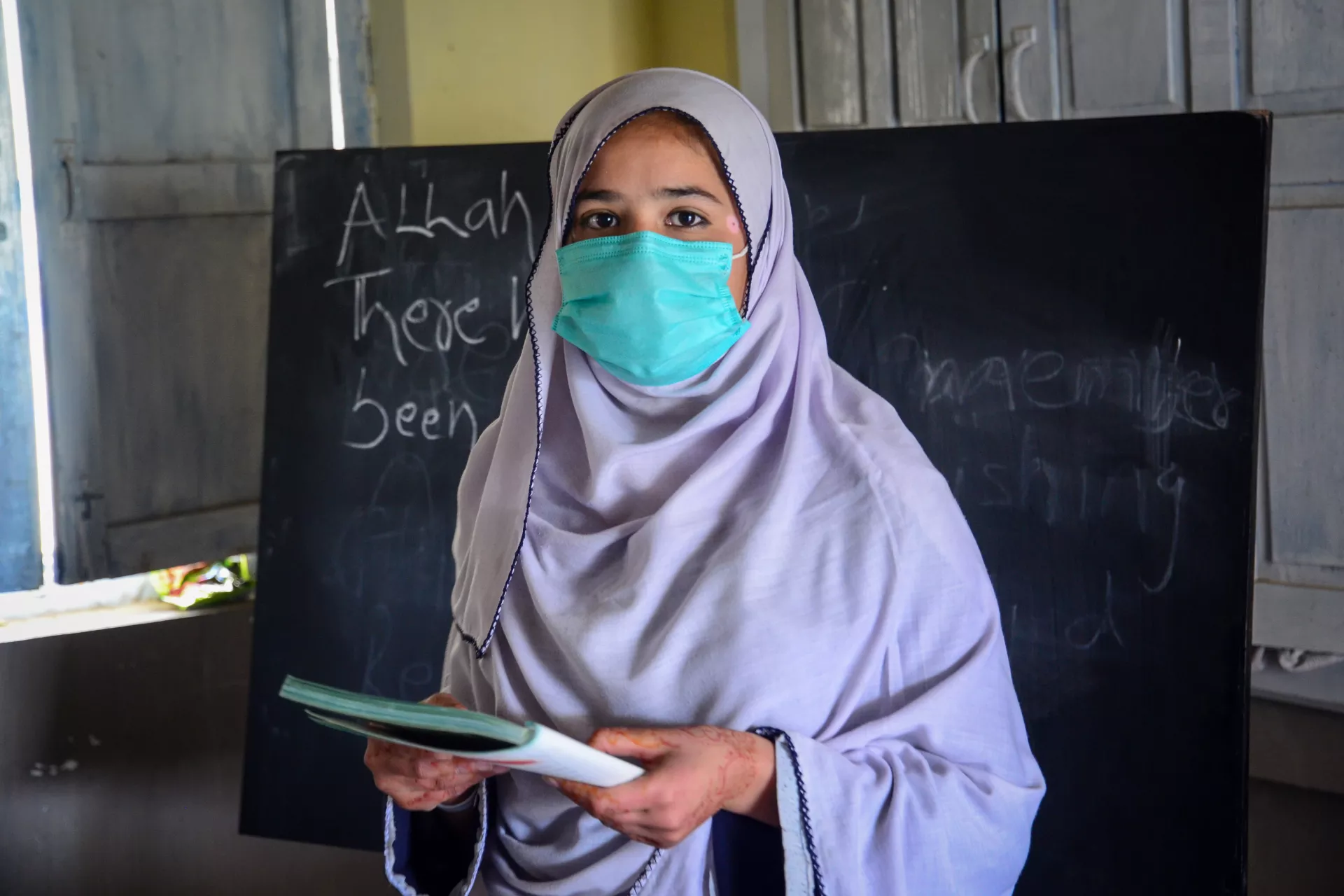 A girl with a facemask on and holding a book stands in front of the blackboard in her classroom in the Hangu area of the Khyber-Pakhtunkhwa province of Pakistan