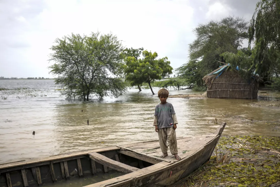 A young boy stands on a wooden boat in a flooded area. The background shows trees partially submerged in water and a small thatched hut on stilts. The sky is overcast, indicating recent heavy rains. The boy looks somber, wearing a gray t-shirt and beige pants, with muddy feet. The scene depicts the impact of flooding in the rural region of Sindh, Pakistan. 