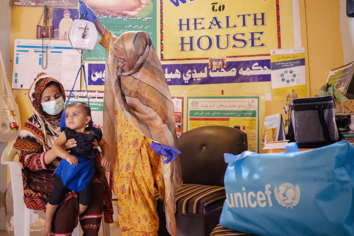Farida uses the infant scale to measure 18-month-old Alisha&rsquo;s weight, as her mother Afshan supports her from below.  