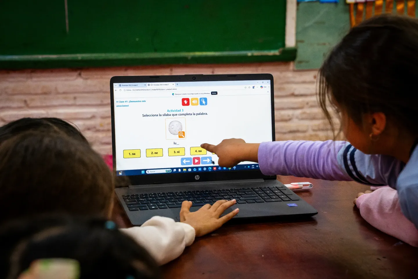 Dos ni&ntilde;as utilizan una computadora port&aacute;til en un aula para realizar una actividad educativa digital de lectura y s&iacute;labas en espa&ntilde;ol.